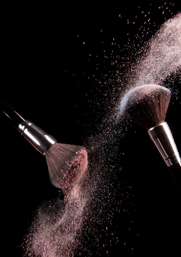 High-speed capture of makeup brushes dispersing pink powder in motion against a dark background.